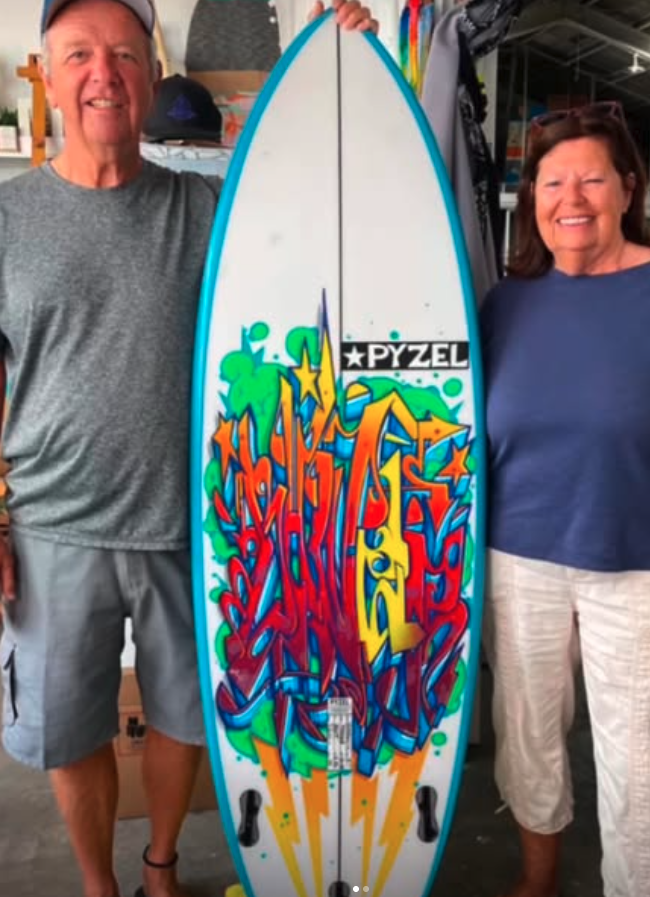A community canvas—surfboard meets street art in a shared creative celebration Vinny and a woman holding a surfboard painted with bold, colorful graffiti