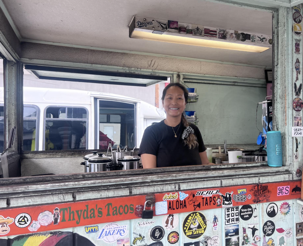 Thyda smiles at the camera from inside the front window of Thyda’s Tacos food truck, surrounded by stickers and kitchen equipment.