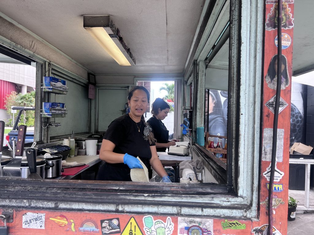 Thyda prepares tacos inside her food truck in Waikiki, wearing a black shirt and blue gloves, with her teammate working behind her.