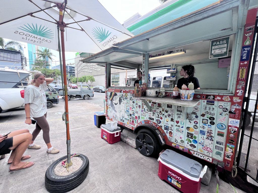 A wide view of Thyda’s Tacos food truck in Waikiki, with customers approaching and the truck covered in colorful stickers under a branded umbrella.
