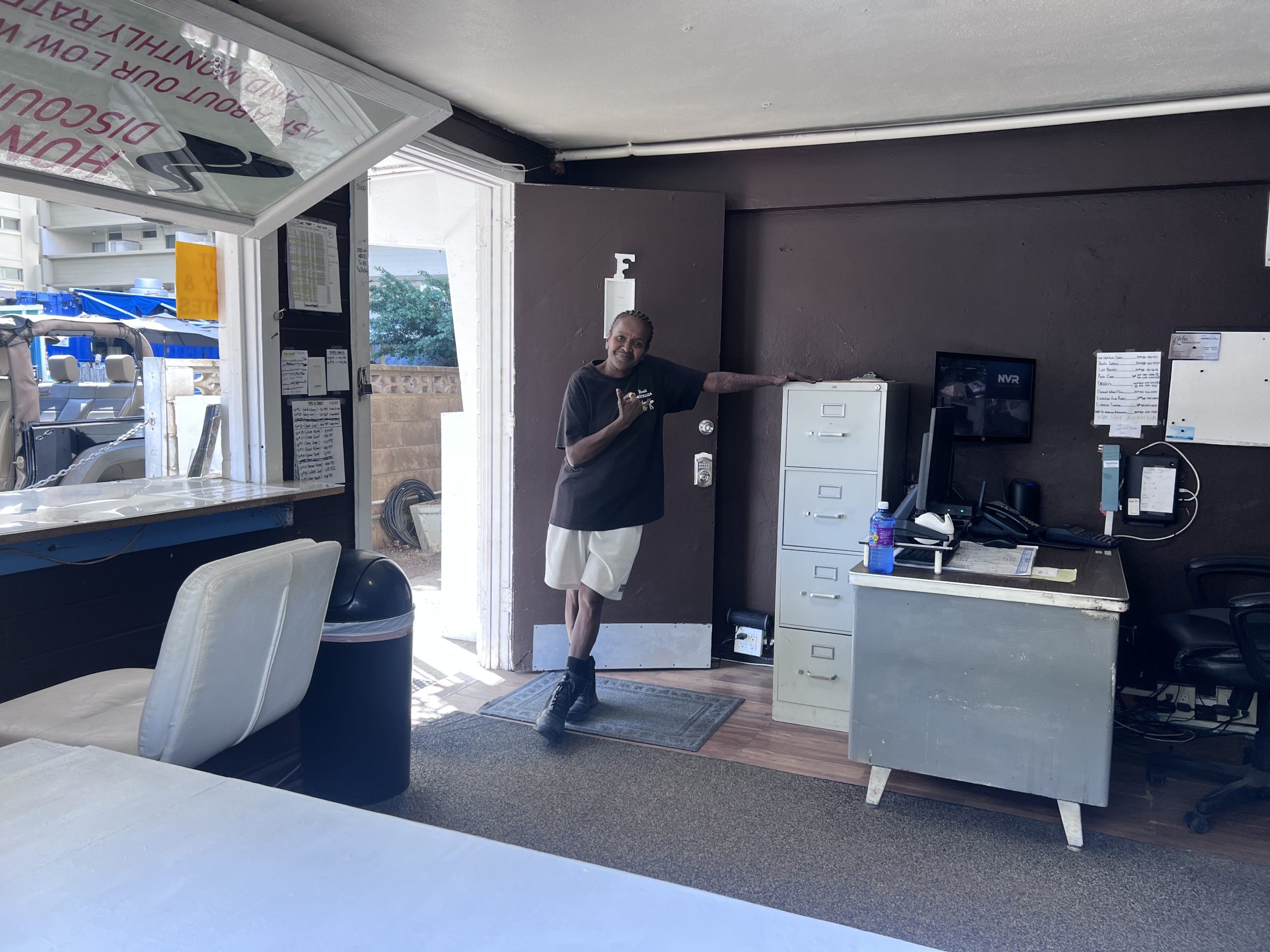 Vicki stands inside the office of Honolulu Discount Cars, leaning casually against a filing cabinet with a warm smile.