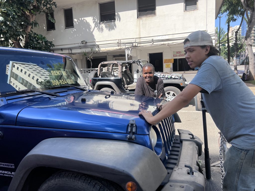 Vicki and a young man stand by a blue Jeep Wrangler parked outside the Honolulu Discount Cars lot, chatting and smiling under the sun.